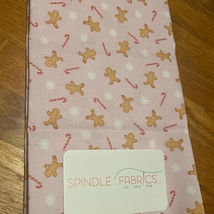 May include: Pink fabric with a repeating pattern of gingerbread men, candy canes, and snowflakes. The fabric has a white border and the text "SPINDLE FABRICS cut sew love" is printed on a white card in the bottom right corner.