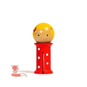 May include: A wooden toy doll with a yellow head, red polka dot pants, and a red bow. The doll is standing on a white surface.