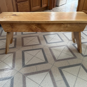 May include: A wooden bench with a natural finish and a simple design. The bench has a rectangular top and two angled legs. The bench is sitting on a tiled floor.