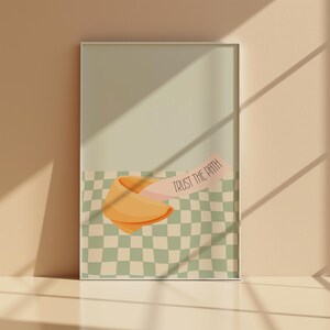 May include: A framed art print featuring a fortune cookie illustration. The fortune cookie is a warm orange color, with a pink fortune that reads "Trust the Path." The background has a checkered pattern in green and white.