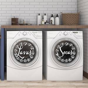 May include: Two white washing machines with black doors. The left door has a black and white wreath with the word "Lavar" in the center. The right door has a black and white wreath with the word "Secar" in the center.