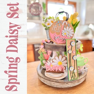 May include: A two-tiered wooden tray decorated with spring-themed wooden cutouts. The top tier features a round wooden sign with a pink umbrella and the text "Hello Spring". The bottom tier features a wooden cutout of a daisy with the text "Welcome" and a wooden sign with a row of daisies and butterflies. The tray is decorated with a string of wooden beads and a small bouquet of artificial daisies and yellow tulips.
