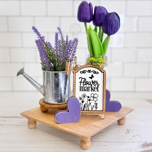 May include: A wooden riser holds a silver metal watering can with purple lavender, a glass jar with three purple tulips, two purple wooden hearts, and a rectangular wooden sign with a scalloped edge that reads Flower market with floral art.