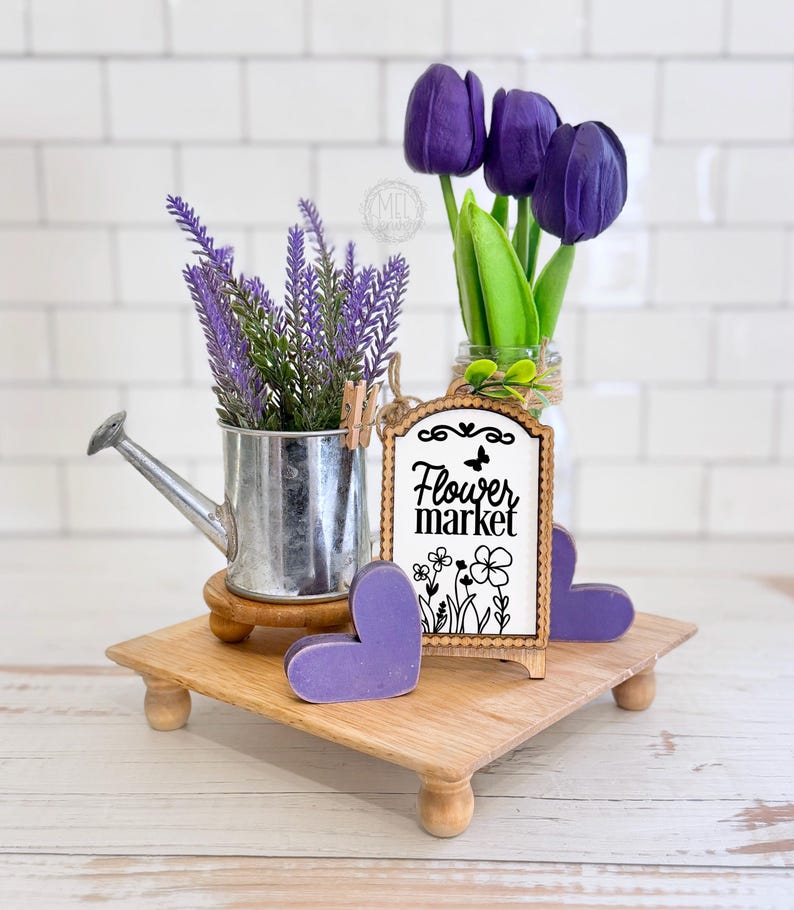 May include: A wooden riser holds a silver metal watering can with purple lavender, a glass jar with three purple tulips, two purple wooden hearts, and a rectangular wooden sign with a scalloped edge that reads Flower market with floral art.