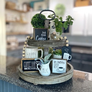May include: A tiered wooden tray with a variety of coffee-themed decorations, including a sign that says "but first coffee", a gnome with a coffee cup hat, a mug that says "happy", and a sign that says "coffee bar open 24 hours".