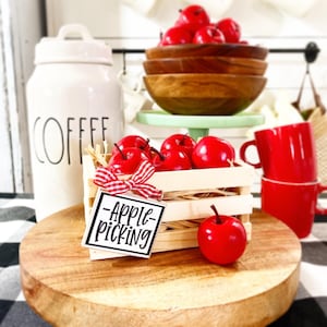 May include: A wooden crate filled with red apples, with a small sign that says "Apple Picking" in black lettering on a white background. The crate is sitting on a wooden surface with a red and black checked pattern.
