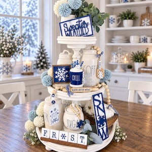 May include: A three-tiered white wooden tray decorated with winter-themed items including a blue sign reading Sweater Weather, a mug labelled Chill, a rolling pin with Warm Winter Wishes, a sign reading SNOW, and tiles spelling out FROST.