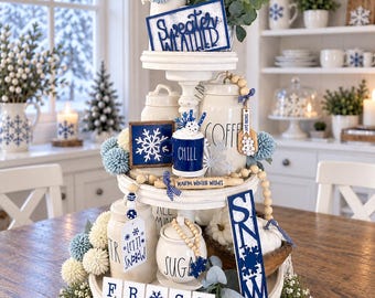 May include: A three-tiered white wooden tray decorated with winter-themed items including a blue sign reading Sweater Weather, a mug labeled Chill, a rolling pin with Warm Winter Wishes, a sign reading SNOW, and tiles spelling out FROST.