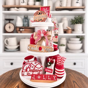 May include: A two-tiered white wooden tray decorated for Christmas with red and white accents. Items include a sign reading Farm Fresh Christmas Trees, a sign reading Caution Sled Parking, a Believe sign, red and white striped straws, and red letter blocks spelling JOLLY.