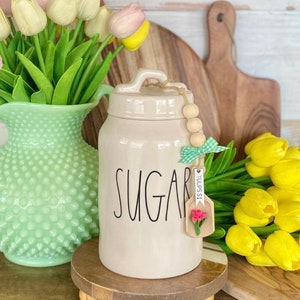 May include: A cream-colored ceramic sugar jar with the word "SUGAR" in black script. The jar has a lid and a wooden tag with a flower and the text "TULIPS $1". The jar sits on a wooden stand, with a green vase of tulips to the left and yellow tulips to the right.
