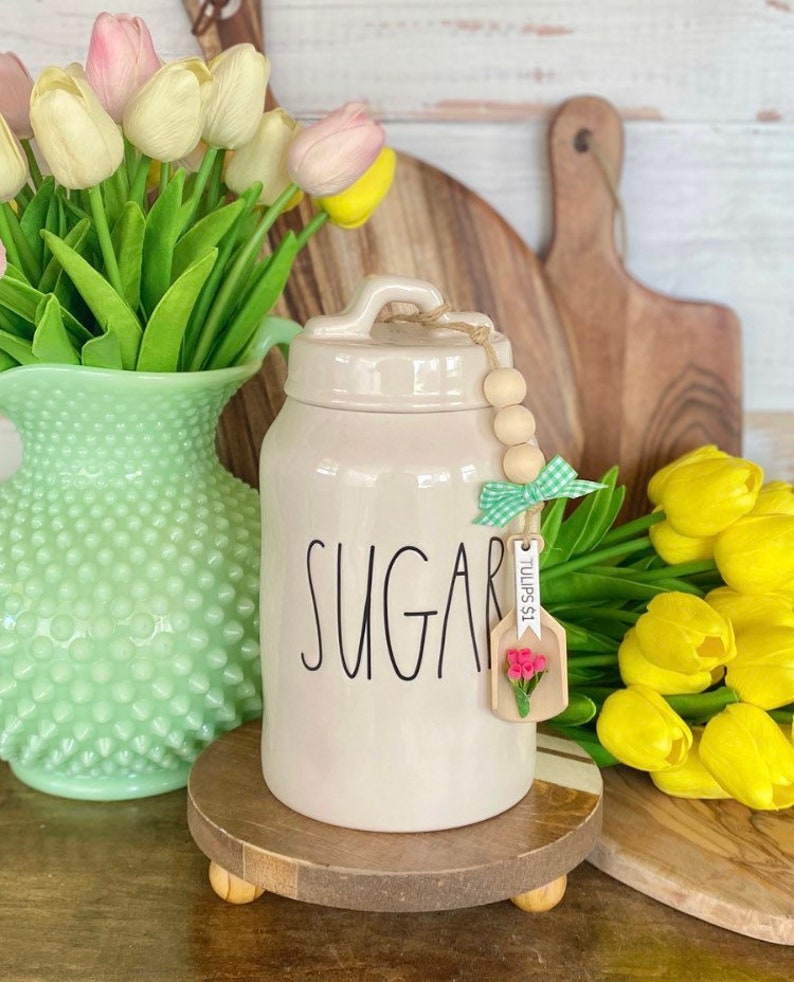 May include: A cream-colored ceramic sugar jar with the word "SUGAR" in black script. The jar has a lid and a wooden tag with a flower and the text "TULIPS $1". The jar sits on a wooden stand, with a green vase of tulips to the left and yellow tulips to the right.