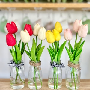 May include: Six glass milk bottles with black and white checkered ribbon tied around the neck, each filled with water and a bouquet of artificial tulips. The tulips are red, white, yellow, and pink.