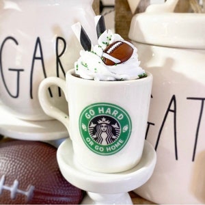Op de afbeelding: Een witte keramische mok met een groen en zwart Starbucks-logo met de tekst "Go Hard or Go Home". De mok heeft slagroom en een miniatuur voetbal erop.