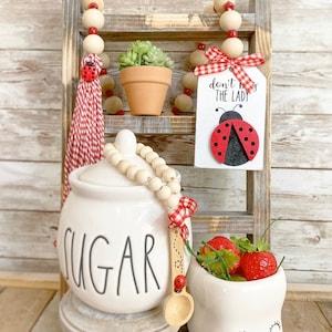 May include: A white ceramic sugar jar with the word "SUGAR" in black lettering. The jar is decorated with a wooden bead garland and a red and white gingham ribbon. A wooden spoon with a ladybug design is next to the jar. A white ceramic mug with a smiling face and red strawberries inside is on the right. A wooden ladder shelf with a sign that reads "Don't Bug The Lady" with a ladybug design is in the background.