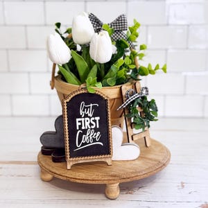 May include: A round wooden tiered tray holding a bucket of white tulips with green leaves and a black and white gingham bow. A small arched chalkboard sign reads but FIRST Coffee in white text, alongside two wooden hearts and a miniature ladder.