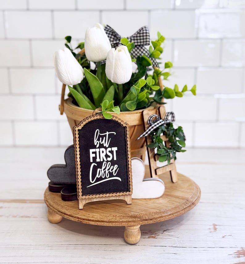 May include: A round wooden tiered tray holding a bucket of white tulips with green leaves and a black and white gingham bow. A small arched chalkboard sign reads but FIRST Coffee in white text, alongside two wooden hearts and a miniature ladder.