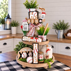 May include: A two-tiered wooden tray decorated with baseball-themed items including signs that read LOVE, WIN, RUN, BALLPARK, GAME DAY, and HOME. The display features miniature baseballs, a wooden bat, artificial greenery, and white ceramic jars.