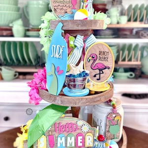 Puede incluir: Una bandeja de madera de tres niveles decorada con artículos con temática de verano. Incluye una tabla de surf, un letrero que dice "SURF", un letrero con un flamenco y un letrero que dice "Hello SUMMER". Otros artículos incluyen pelotas de playa y un paraguas pequeño.