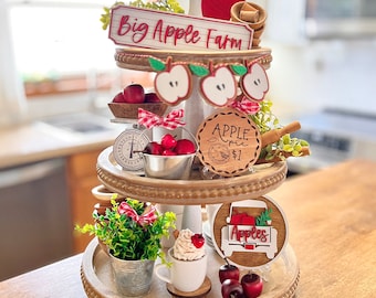 Puede incluir: Una bandeja de madera de tres niveles decorada con artículos de temática otoñal, incluyendo manzanas rojas, un letrero de madera que dice "Big Apple Farm", un letrero de madera que dice "Apple Pie 1€", y un letrero de madera que dice "Apples".