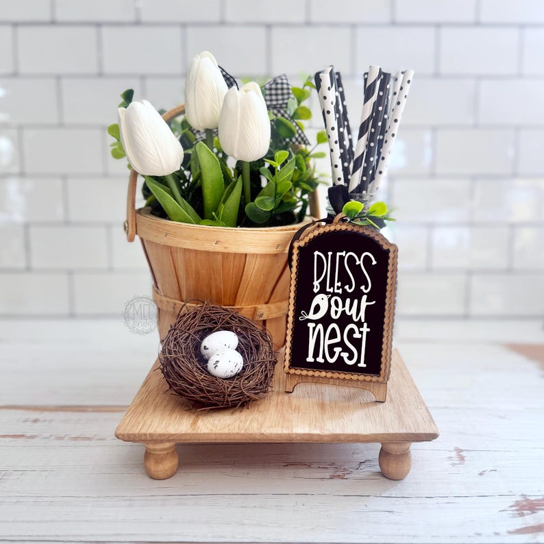 May include: A wooden basket filled with white tulips and green leaves, a small bird's nest with two speckled white eggs, and a black arched sign with white text that reads Bless Our Nest, accompanied by black and white patterned paper straws.