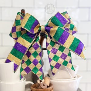 May include: A large decorative bow with a Mardi Gras theme. The bow features a gold, green, and purple diamond pattern. The bow is made of a glittery ribbon and is displayed on a tiered tray with white bowls and wooden spools.