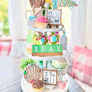May include: A three-tiered white tiered tray decorated with a birthday theme. The top tier has a wooden sign that says "Party", a wooden sign that says "Life is better with sprinkles", a cupcake, and a wooden mug with a beaded garland. The middle tier has a green and white striped ribbon, a green and white checkered ribbon, a green and white striped box, and wooden blocks that spell out "B.D.A.Y." The bottom tier has a wooden sign that says "Happy Birthday", a wooden sign that says "April 14", a cupcake, and a glass milk bottle with a pink drink.