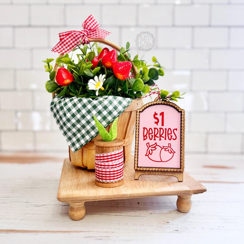 May include: A wooden basket filled with artificial greenery, red strawberries, and a white daisy, accented by a red and white gingham bow. Beside it sits a wooden spool with red ribbon and a pink sign with the text $1 BERRIES and strawberry icons.