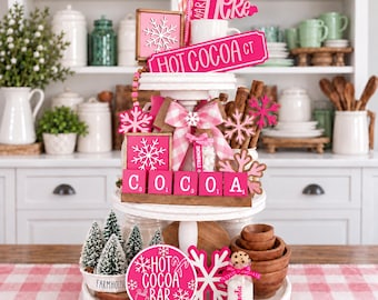 May include: A white three-tiered tray decorated with pink and white winter-themed items, including signs reading HOT COCOA CT, WARM UP HERE, and HOT COCOA BAR, pink snowflake cutouts, a pink gingham bow, mini trees, and a small jar labeled Santa.