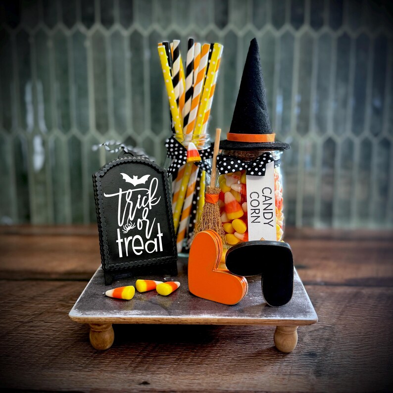 May include: A Halloween tiered tray decor set featuring a black sign with white text reading Trick or Treat, a glass jar filled with candy corn topped with a black witch hat, a small broom, orange and black heart shapes, and paper straws.