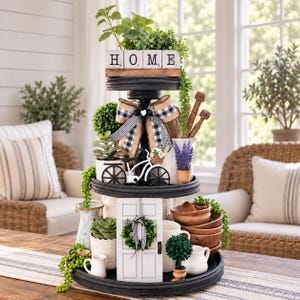 May include: A two-tiered black wooden tray decorated with various items including white blocks spelling HOME, a black and white buffalo check bow, a white bicycle figurine, a miniature white door with a wreath, wooden bowls, and faux greenery.