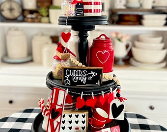 May include: A three-tiered tiered tray decorated for Valentine's Day with red and white hearts, a red cardinal, a sign that says "Cupid's Candy Co.", and a red and white striped jar with red flowers.
