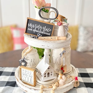 Könnte beinhalten: Ein zweistufiges, weißes, dekoratives Tablett mit verschiedenen religiösen Gegenständen. Enthält eine Tafel mit dem Text "amazing grace", eine kleine Kirche, Engelsfiguren und eine Teigrolle mit dem Text "faith family love". Rosa Tulpen in einer weißen Vase.