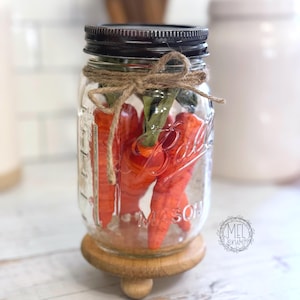 May include: A clear Mason jar with a black lid, filled with orange carrots and green stems, tied with twine. The jar sits on a wooden riser. The jar is 5.25 inches tall and 2.75 inches wide.
