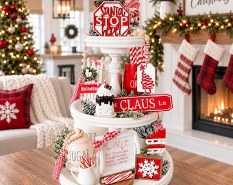 Könnte beinhalten: Ein weißes, dreistöckiges Holztablett, weihnachtlich dekoriert mit Schildern wie Santa Stop Here, Claus Ln und Mrs. Claus Cafe. Das Arrangement enthält ein Zuckergefäß, einen Becher mit Zuckerstangen, ein Schneeflockenschild und kleine Schlitten.