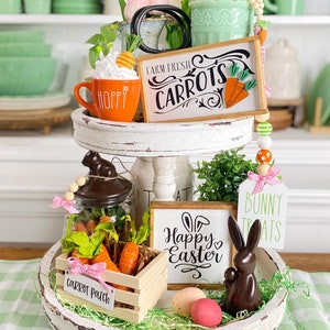 Pode incluir: Uma bandeja branca de dois níveis decorada para a Páscoa. O nível superior apresenta uma caneca com a palavra "Hoppy" nela, uma placa que diz "Farm Fresh Carrots" e um pequeno buquê de tulipas rosa. O nível inferior apresenta uma caixa de madeira com as palavras "Carrot Patch" nela, um frasco de vidro com cenouras, uma placa que diz "Happy Easter", um coelho de chocolate e ovos de Páscoa.
