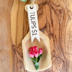 May include: A wooden scoop-shaped ornament with a small bouquet of pink tulips. The ornament has a white tag that reads "TULIPS $1" and is suspended by a twine string and a green and white checkered ribbon.