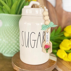 May include: Cream-colored ceramic sugar jar with the word "SUGAR" in black script. The jar has a lid with a handle and a wooden bead and ribbon accent. A small wooden tag with pink flowers hangs from the jar.