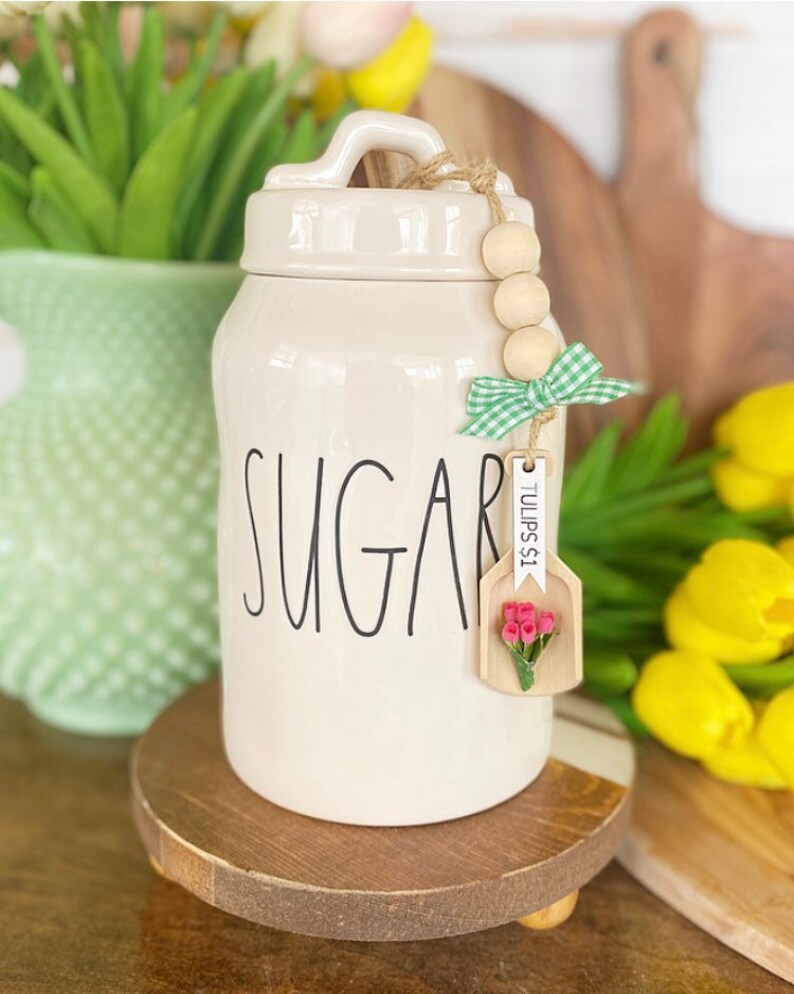 May include: Cream-colored ceramic sugar jar with the word "SUGAR" in black script. The jar has a lid with a handle and a wooden bead and ribbon accent. A small wooden tag with pink flowers hangs from the jar.