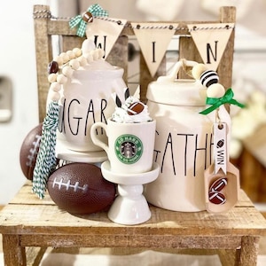 May include: White ceramic canisters with black lettering that reads "Sugar" and "Gather". The canisters are decorated with wooden bead garlands and green and white striped ribbon. A small white pedestal holds a coffee mug with a green Starbucks logo that reads "Go Hard or Go Home". The mug is topped with whipped cream and a miniature football. A wooden sign with the word "WIN" is hanging on the side of the canisters.