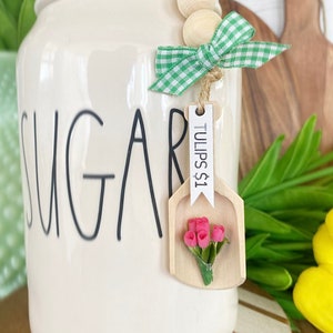 May include: A cream-colored sugar jar with the word "SUGAR" in black script. A wooden scoop-shaped tag with "TULIPS $1" and a small bouquet of pink flowers hangs from a green and white gingham bow.