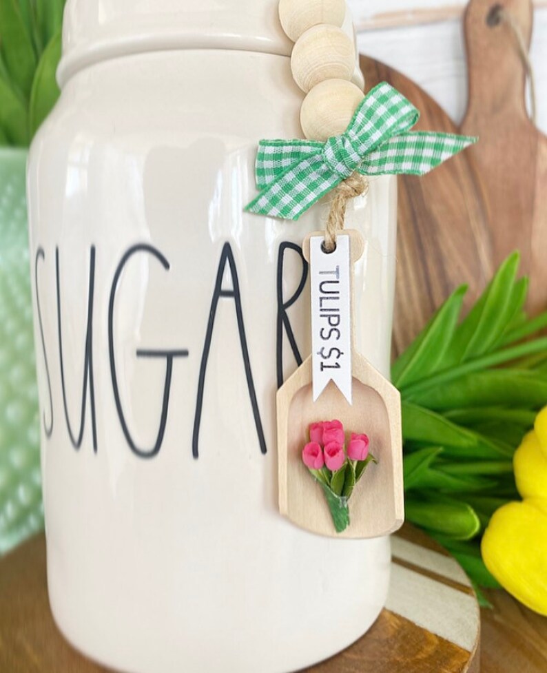 May include: A cream-colored sugar jar with the word "SUGAR" in black script. A wooden scoop-shaped tag with "TULIPS $1" and a small bouquet of pink flowers hangs from a green and white gingham bow.