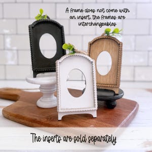 May include: Three arched picture frames in black, white, and natural wood, each featuring a scalloped edge and a central oval cutout. Each frame is topped with a small green leaf accent. Text reads: A frame does not come with an insert. The frames are interchangeable. The inserts are sold separately.