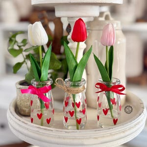 May include: Three glass milk bottles with red hearts printed on them, each holding a single tulip. The tulips are white, pink, and red.