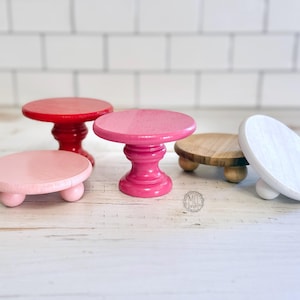 May include: Four wooden cake stands in different sizes and colors. The stands are painted pink, red, and white. The stands are perfect for displaying cakes, cupcakes, or other treats.