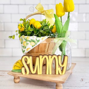 May include: A wooden tray holds a decorative arrangement. A basket filled with lemons and greenery sits next to a glass jar with yellow tulips. The word "SUNNY" is displayed in yellow and brown wooden letters, with a lemon slice on the side.