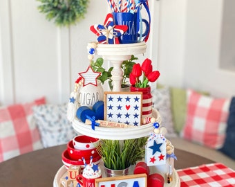 Pode incluir: Uma bandeja branca de três níveis decorada com itens temáticos patrióticos. O nível superior apresenta uma caneca azul com "USA", juntamente com decorações em forma de estrela e canudos vermelhos, brancos e azuis. O nível do meio tem uma placa que diz "Miss American Pie".