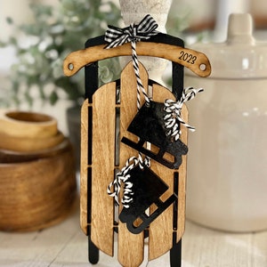 May include: Wooden sled ornament with black accents and two black ice skate cutouts. The sled has a black and white checkered bow and the year "2023" etched on the side. The sled is made of light brown wood.
