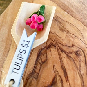 May include: A small wooden scoop holds a bouquet of pink tulips with green leaves. A white tag with the text "TULIPS $1" hangs from the scoop. The background is a wooden surface with a natural wood grain.