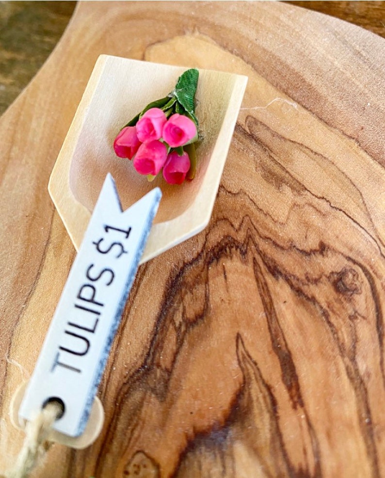 May include: A small wooden scoop holds a bouquet of pink tulips with green leaves. A white tag with the text "TULIPS $1" hangs from the scoop. The background is a wooden surface with a natural wood grain.