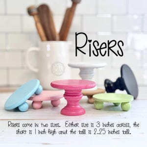 May include: A collection of small, colorful wooden risers in various shades including pink, blue, green, and gray. The risers are round with three or four ball feet. Text on the image reads "Risers" and measurements: 3 inches across, 1 inch high, and 2.25 inches tall.